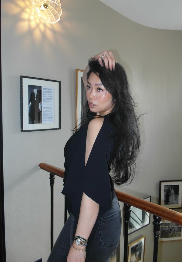 Femme aux longs cheveux noirs portant un haut noir à épaules dénudées et un jean, posant sur un escalier intérieur avec des photos encadrées et une lampe murale allumée au-dessus.