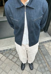 Persoon in een donker denimjack over een witte blouse, lichte beige broek en zwarte tassel loafers, staand op betegelde stoep.