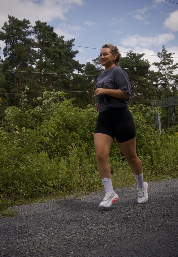 Femme qui fait du jogging sur un chemin de gravier à côté de buissons verts et d’arbres sous un ciel partiellement nuageux.