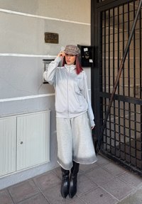 Žena s červenými vlasmi, oblečená v sivej kockovanej šiltovke, svetlosivej bundě Adidas, širokých sivých nohaviciach a čiernych špicatých čižmách, stojaca pri kovovej bráne.