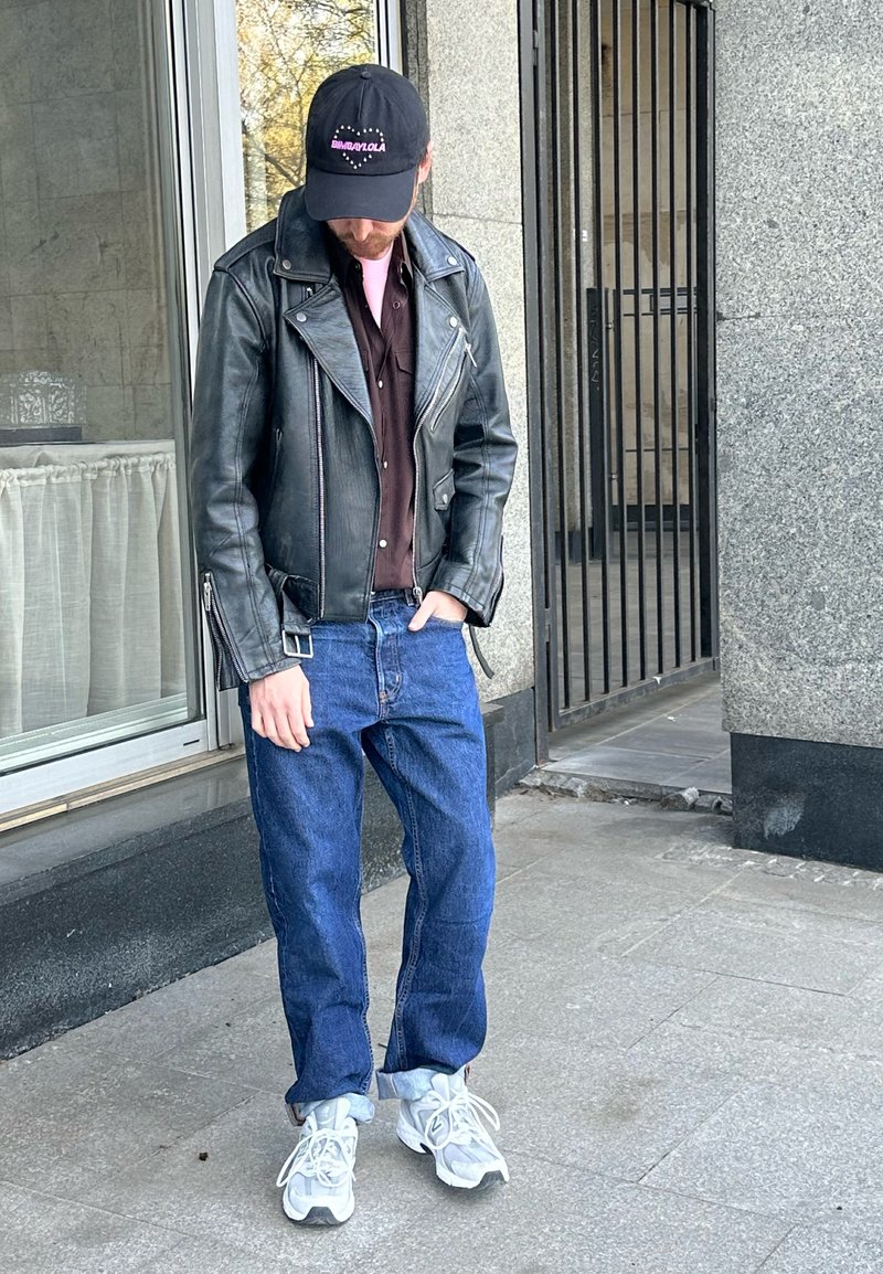 Homme portant une veste en cuir noire, une chemise foncée, un jean bleu, des baskets blanches et une casquette noire, debout sur le trottoir près de l'entrée du bâtiment.