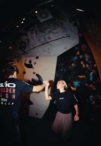 Due persone si danno un cinque in una palestra di arrampicata con una parete texturizzata e prese colorate. Entrambi indossano magliette nere con un logo e dettagli sull'evento.