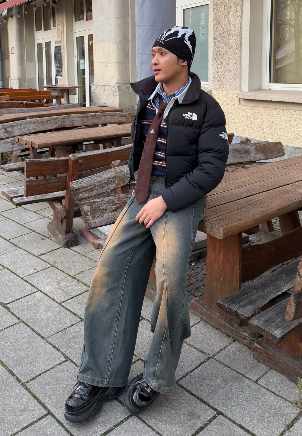 Chaqueta de abrigo negra, camisa a rayas, corbata de estampado, jeans de pierna ancha y desgastados, y zapatos de plataforma negros brillantes, sentado en bancos de madera al aire libre.