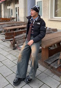 Chaqueta de abrigo negra, camisa a rayas, corbata de estampado, jeans de pierna ancha y desgastados, y zapatos de plataforma negros brillantes, sentado en bancos de madera al aire libre.