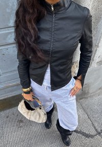 Jaqueta de couro preta com colarinho elevado, fecho de zip, combinada com calças de moletom cinza claro. Segurando uma bolsa de cor clara e usando botas pretas.