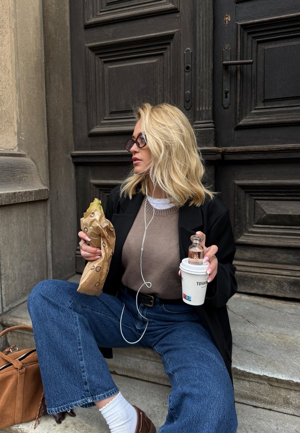 Blond woman wearing glasses and a dark blazer, sitting on stone steps, holding a wrapped sandwich and a coffee cup with a perfume bottle on top.