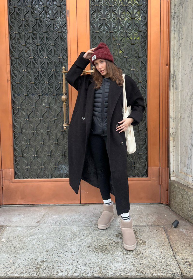 Manteau en laine noire porté sur une doudoune noire, accompagné de leggings noirs et de bottines beiges. Un bonnet tricoté bordeaux et un sac fourre-tout crème complètent la tenue.