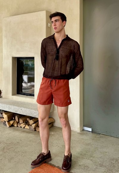 Camisa negra traslúcida, pantalones cortos de baño naranjas y zapatos marrón oscuro. Pose individual con un fondo neutro y una chimenea. Elementos de diseño simples.