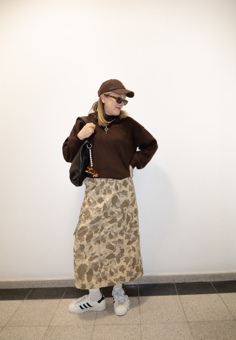 Femme portant une casquette marron, des lunettes de soleil et un pull, avec une jupe camouflage beige et des baskets blanches, portant un sac noir, debout à l'intérieur sur un sol carrelé.