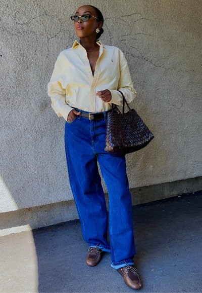Camisa amarilla de botones, jeans de tiro alto azules, bolso trenzado marrón, zapatos marrones, gafas de sol, contra una pared texturizada neutra.