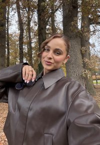 Brun oversizejacka i konstläder med bred krage och stora ärmar. Ett par solglasögon hålls i ena handen.