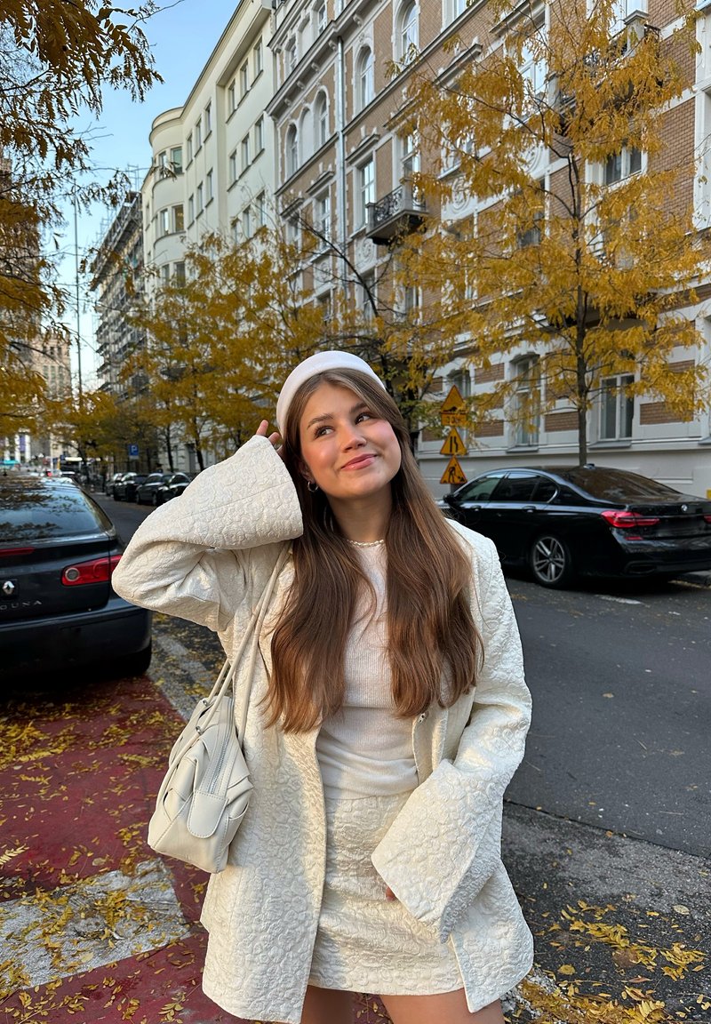 Cappotto trapuntato chiaro e testurizzato, top bianco e gonna. Tiene una piccola borsa chiara. Scenario urbano con foglie autunnali e auto parcheggiate.