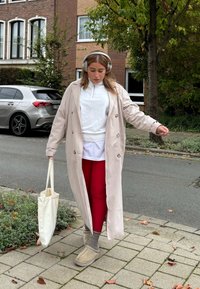 Lång beige kappa, vit topp, röda leggings, grå strumpor och beige tofflor. Håller en krämfärgad tote-väska, går på en stenbelagd gång.