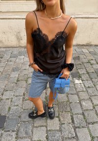 Fekete csipkével díszített camisole, kék mini táska vállpánttal, farmer rövidnadrág és fekete loaferek fém részletekkel kövezet utcán.