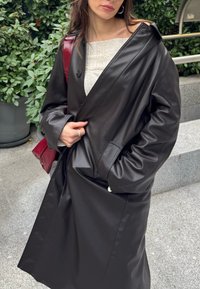 Femme en plein air portant un long manteau en cuir noir sur un haut beige clair, portant un sac à main rouge, une main dans la poche et l'autre sur le manteau.