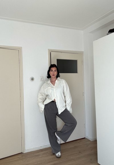 Mujer de pie sobre una pierna en una habitación, vestida con una camisa blanca de gran tamaño, pantalones grises y zapatillas blancas, con las manos en los bolsillos.