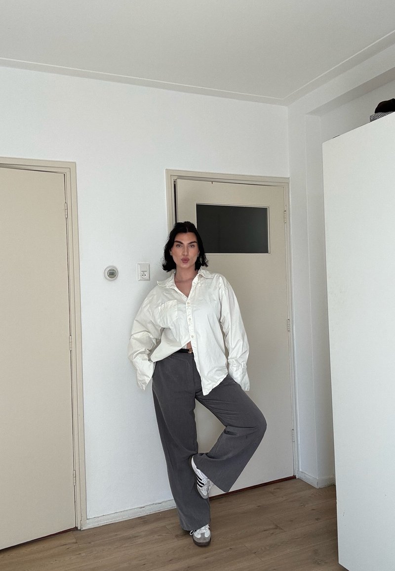 Femme debout sur une jambe dans une pièce, portant une chemise blanche surdimensionnée, un pantalon gris et des baskets blanches, les mains dans les poches.