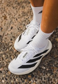 Sapatilhas brancas de corrida com riscas diagonais pretas, parte superior em malha texturizada e atacadores planos, combinadas com meias brancas caneladas com um logo preto.