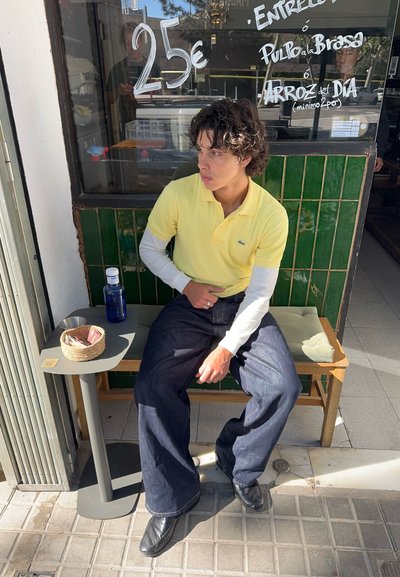 Joven con polo amarillo y vaqueros oscuros sentado en un banco fuera de una cafetería con pared de azulejos verdes y menú manuscrito en la ventana.