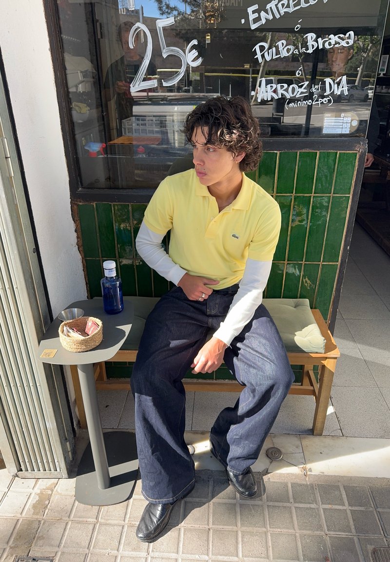 Young person in yellow polo and dark jeans sitting on bench outside cafe with green tiled wall and handwritten menu on window.
