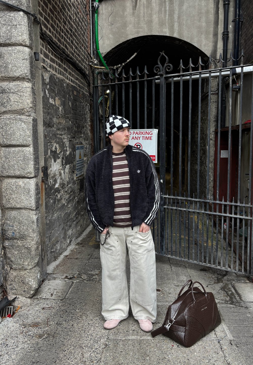 Jeune homme en chemise rayée et chapeau à carreaux, debout près d'une arche avec portail, avec un grand sac marron posé au sol à côté de lui.