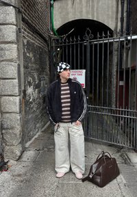 Jeune homme en chemise rayée et chapeau à carreaux, debout près d'une arche avec portail, avec un grand sac marron posé au sol à côté de lui.