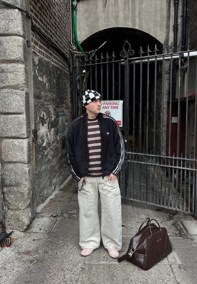 Hombre joven con camiseta de rayas y sombrero a cuadros, de pie junto a un arco con reja, con una gran bolsa marrón en el suelo a su lado.