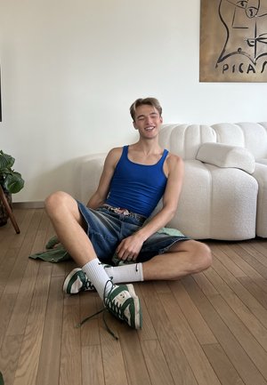 Joven con camiseta sin mangas azul y pantalones cortos de mezclilla sentado con las piernas cruzadas en un suelo de madera, sonriendo con los ojos cerrados, cerca de un sofá blanco curvado.