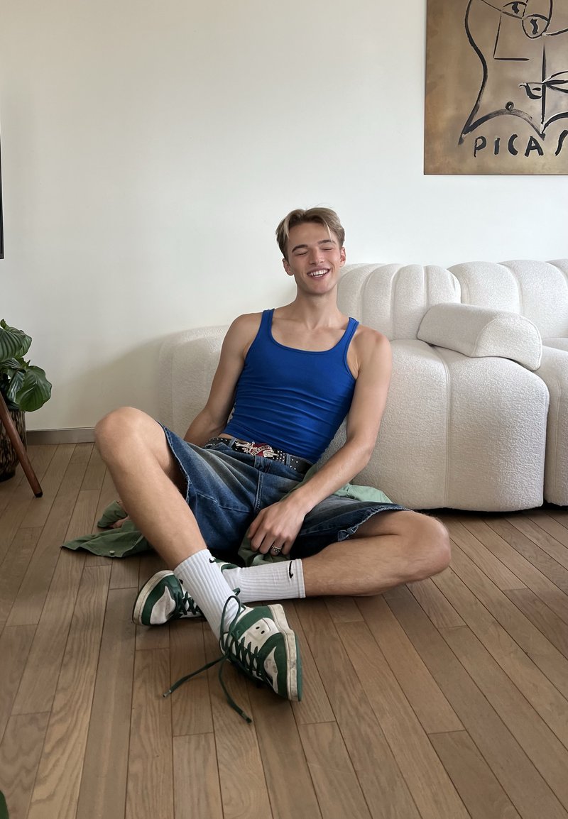 Young man in blue tank top and denim shorts sits cross-legged on wooden floor, smiling with eyes closed, near white curved sofa.