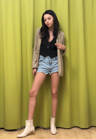 Un blazer a cuadros claro sobre una blusa negra con botones, combinado con pantalones cortos de denim desgastados y botas blancas hasta el tobillo, contra un fondo texturizado verde.
