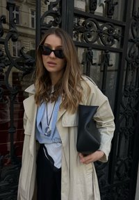 Bež trench coat, svetlo modra srajca, črne hlače in prevelika črna torba. Model nosi temna sončna očala in srebrno ogrlico.