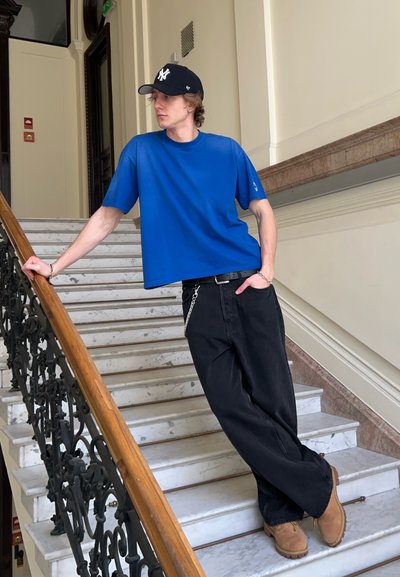 Camiseta azul de manga corta, pantalones negros sueltos, zapatos marrones y gorra negra. De pie en una escalera con peldaños de mármol y barandillas decorativas.