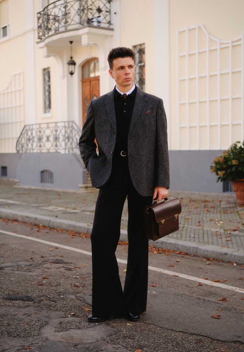 Blazer grigio in tweed sopra camicia nera, pantaloni neri larghi, scarpe lucide, tiene una valigetta in pelle marrone. Dettaglio sobrio del fazzoletto da taschino.