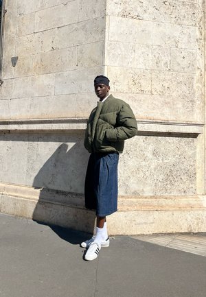 Chaqueta bomber acolchada de color verde oliva, pantalones cortos de mezclilla azul holgados, calcetines blancos y zapatillas de deporte en blanco y negro, de pie junto a una pared de piedra.