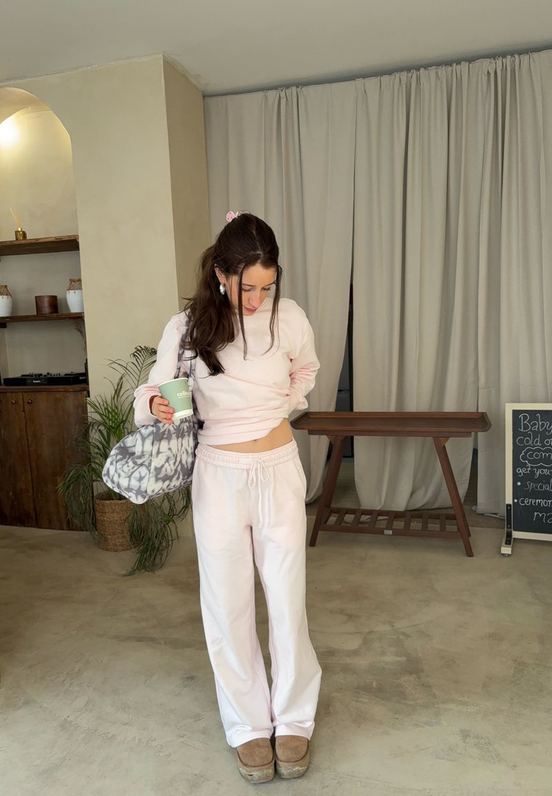 Jeune femme en tenue décontractée rose pastel, tenant une tasse verte et un sac à motifs, debout sur un sol en béton dans un intérieur minimaliste.