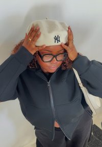 Berretto di lana bianco con un logo nero, indossato da una persona in una felpa nera con zip. Sono visibili pelle chiara, capelli ricci e grandi occhiali.