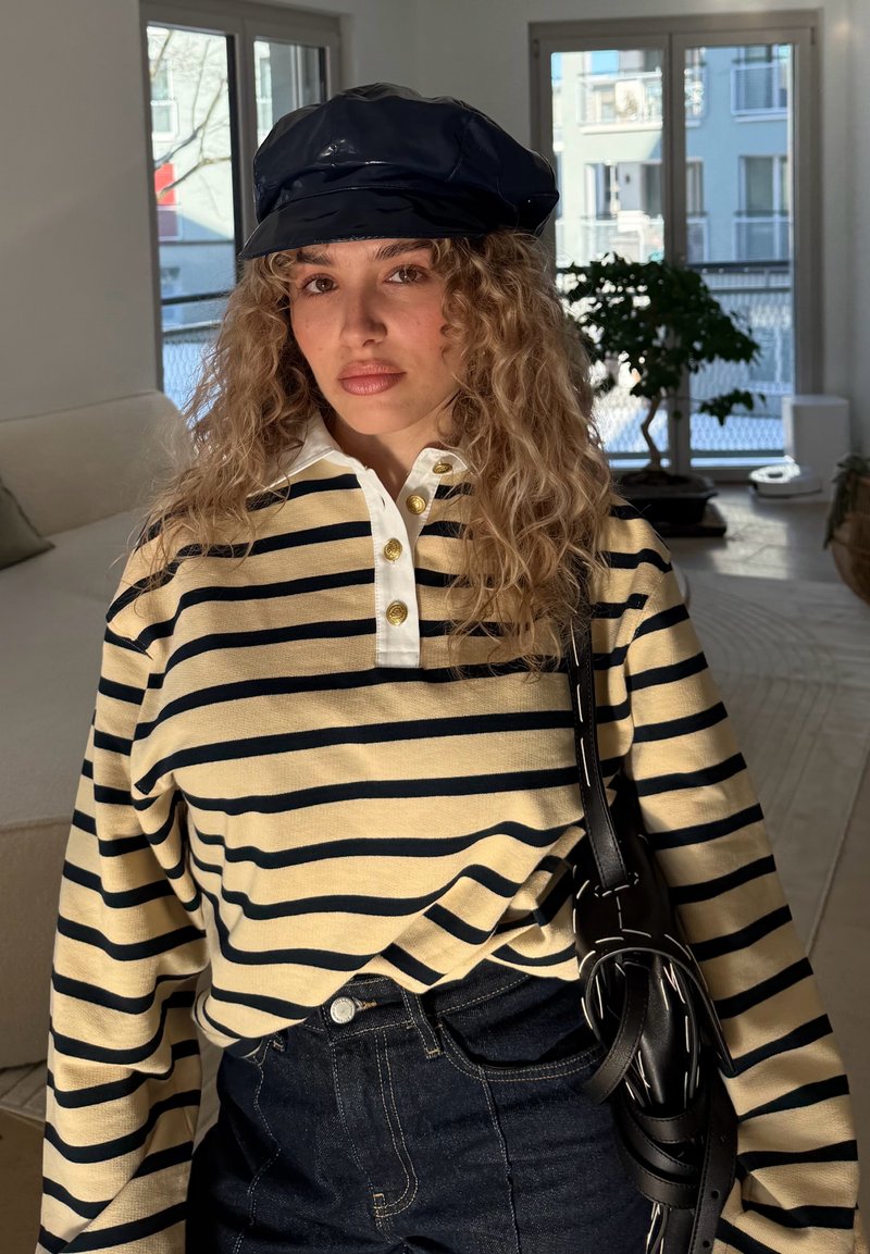 Femme aux cheveux bouclés portant une casquette noire, un t-shirt à manches longues à rayures beige et noire, un jean foncé et un sac à bandoulière noir à l'intérieur.
