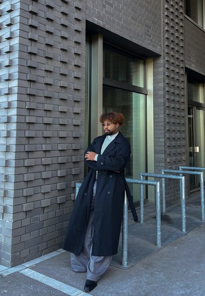 Abrigo trinchera negro, pantalones gris con patrón, cuello alto blanco, de pie contra una pared de ladrillo gris texturizada, con soportes para bicicletas de metal cerca.