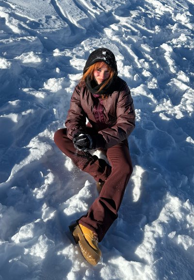 Persona sentada con las piernas cruzadas sobre la nieve, vistiendo una chaqueta marrón, pantalones a juego, capucha negra, guantes y botas color beige, sonriendo a la cámara.