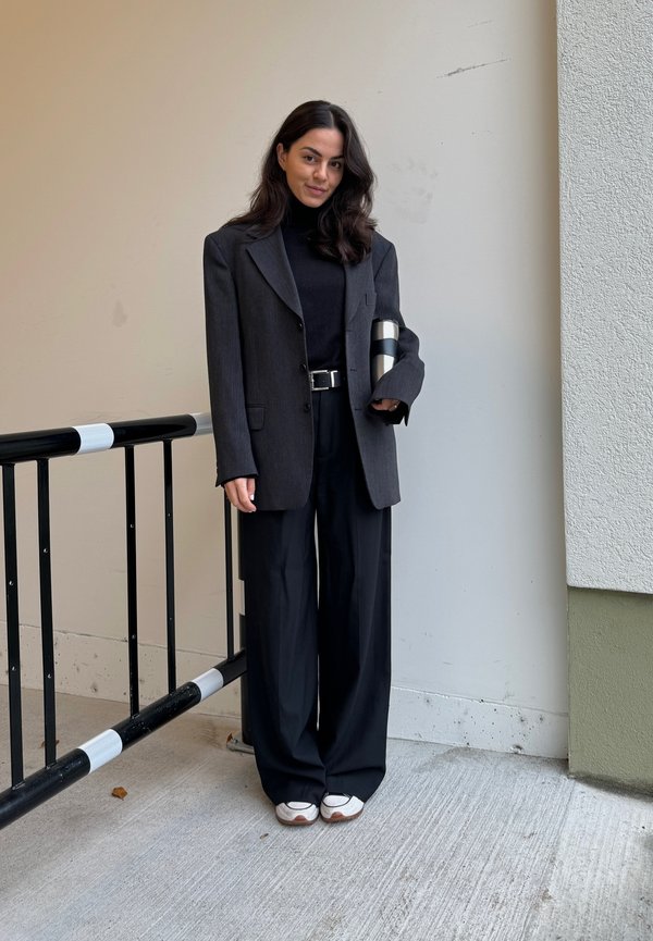 Blazer nero a righe sottili su un dolcevita nero, pantaloni neri a gamba larga e sneaker bianche. Tiene in mano una borraccia termica argentata.