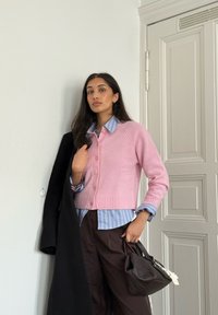 Lichtroze gebreid vest met knopen, gedragen over een lichtblauw gestreept hemd. Donkerbruine broek; houdt een zwarte jas en een bruine handtas vast.