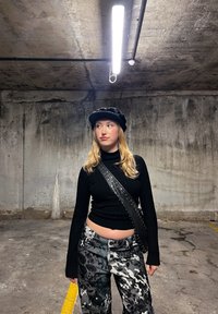 Black ribbed long-sleeve top, paired with textured, patterned gray pants. A black hat and studded crossbody strap complete the look.