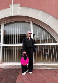 Μαύρη φόρμα, φαρδιά μαύρα παντελόνια, ροζ φουντωτή τσάντα, και ροζ καπέλο με μαντήλι. Τοποθετημένος μπροστά από στρογγυλό παράθυρο με μεταλλικές μπάρες.