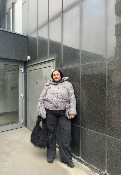 Chaqueta de plumas gris con mangas oversized, pantalones negros y un bolso negro y esponjoso. Superficie pavimentada y pared oscura y reflectante en el fondo.