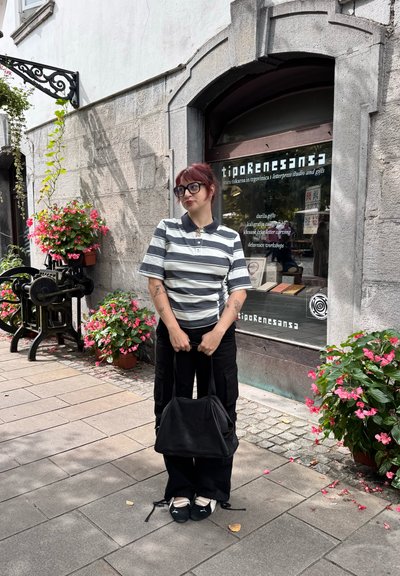 Camiseta polo de manga corta a rayas en gris y blanco, pantalones cargo negros, sosteniendo un bolso negro, con plantas y flores de fondo.