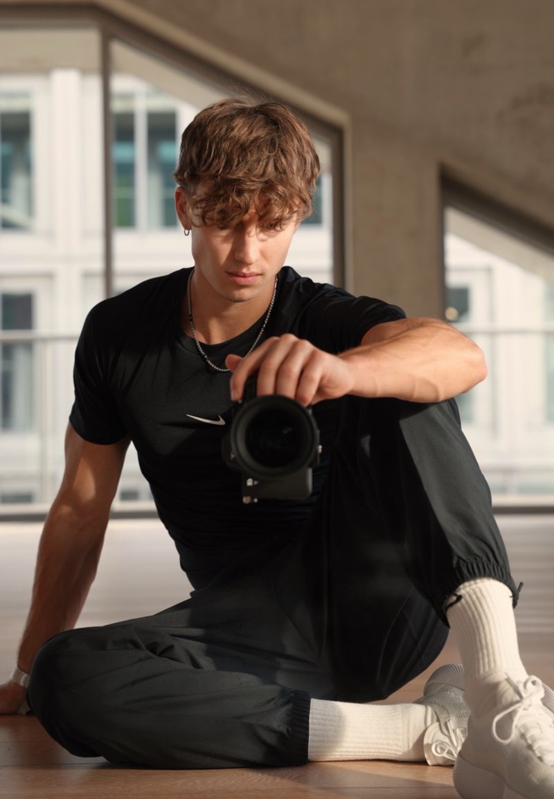 Svart Nike-t-shirt, svarta joggers med elastiska muddar, vita strumpor och vita sneakers. Personen håller i en kamera och sitter på ett trägolv.