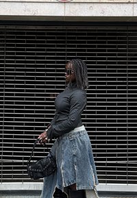 Čierna priliehavá košeľa s dlhým rukávom, rifľová sukňa s asymetrickým lemom a strapcami, čierna kabelka so cvokmi, slnečné okuliare, textúrované vlasy.