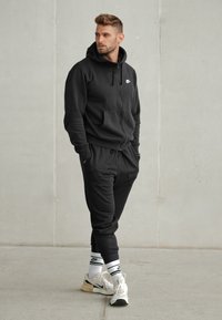 Hanorac negru cu fermoar și pantaloni din fleece asortați, având un buzunar frontal și logo-ul alb Nike. Asortați cu adidași albi și negri.