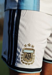 Shorts de sport blancs avec des rayures noires et un emblème doré comprenant trois étoiles et un détail de drapeau bleu et blanc.