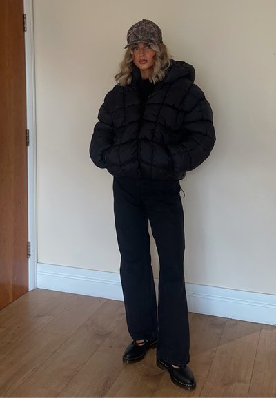 Chaqueta acolchada negra con un acabado brillante, de corte oversize y capucha, combinada con pantalones de campana negros y zapatos negros. Gorra de camuflaje.